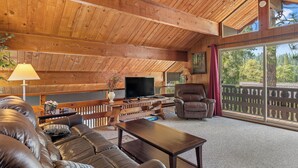 A spacious living area with a wooden ceiling, leather sofas, a television, and a large window with a view of trees.