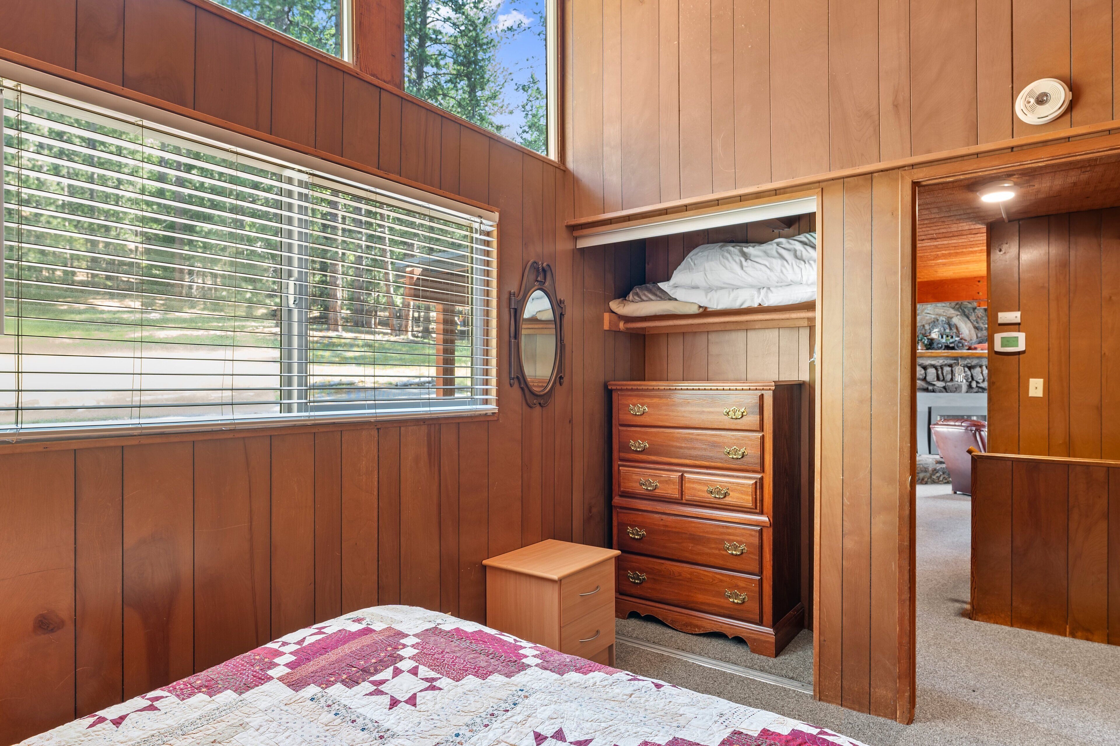 A bedroom with a wooden paneled wall, a bed with a quilt, a dresser, and a window with blinds.