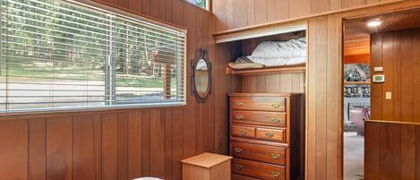 A bedroom with a wooden paneled wall, a bed with a quilt, a dresser, and a window with blinds.