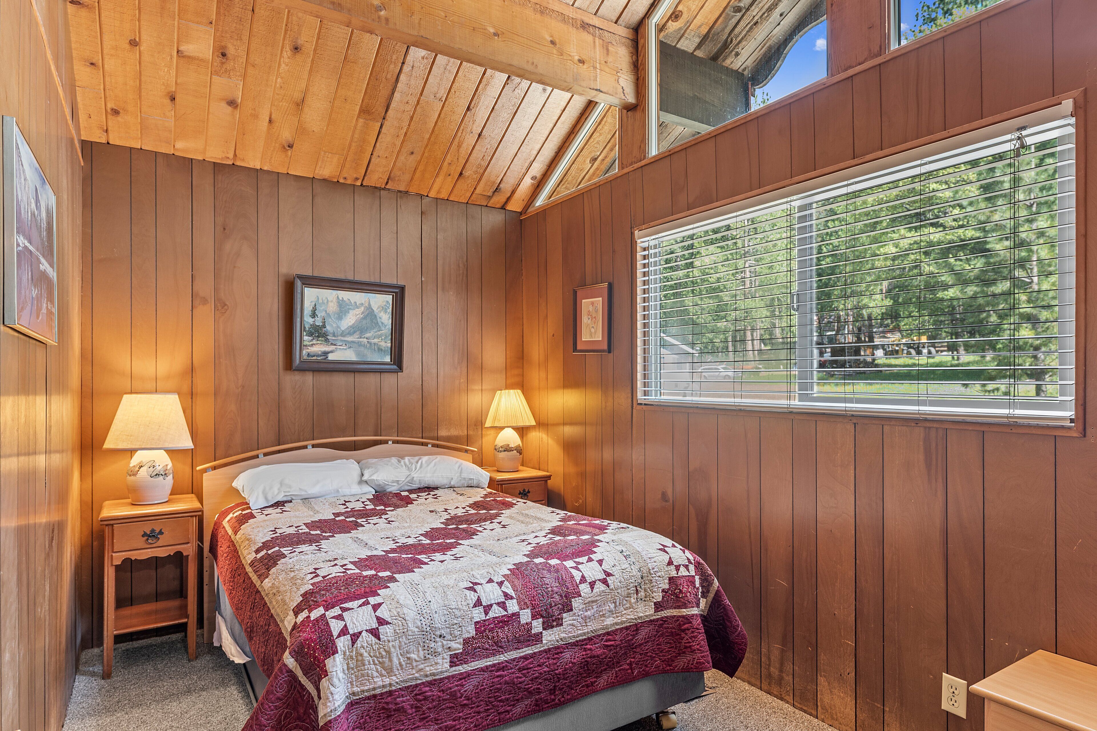 Un dormitorio con paneles de madera, una cama grande, dos lámparas de noche, un cuadro enmarcado en la pared y una ventana con vistas a árboles.
