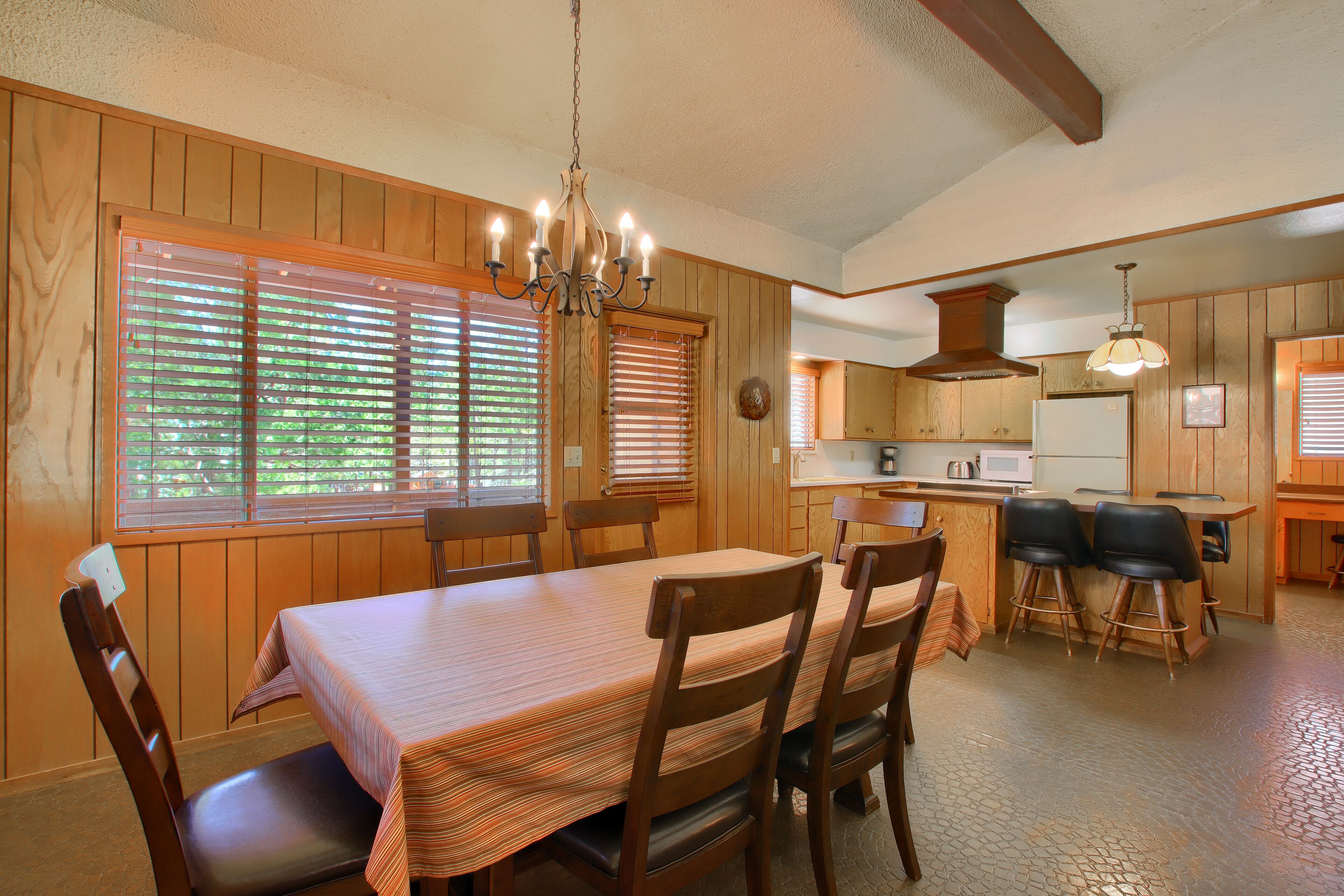 A dining area with a wooden table and chairs, a chandelier, and a kitchen in the background.