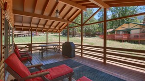 A covered outdoor seating area with wooden benches and a table, overlooking a grassy landscape with trees and a building in the background.