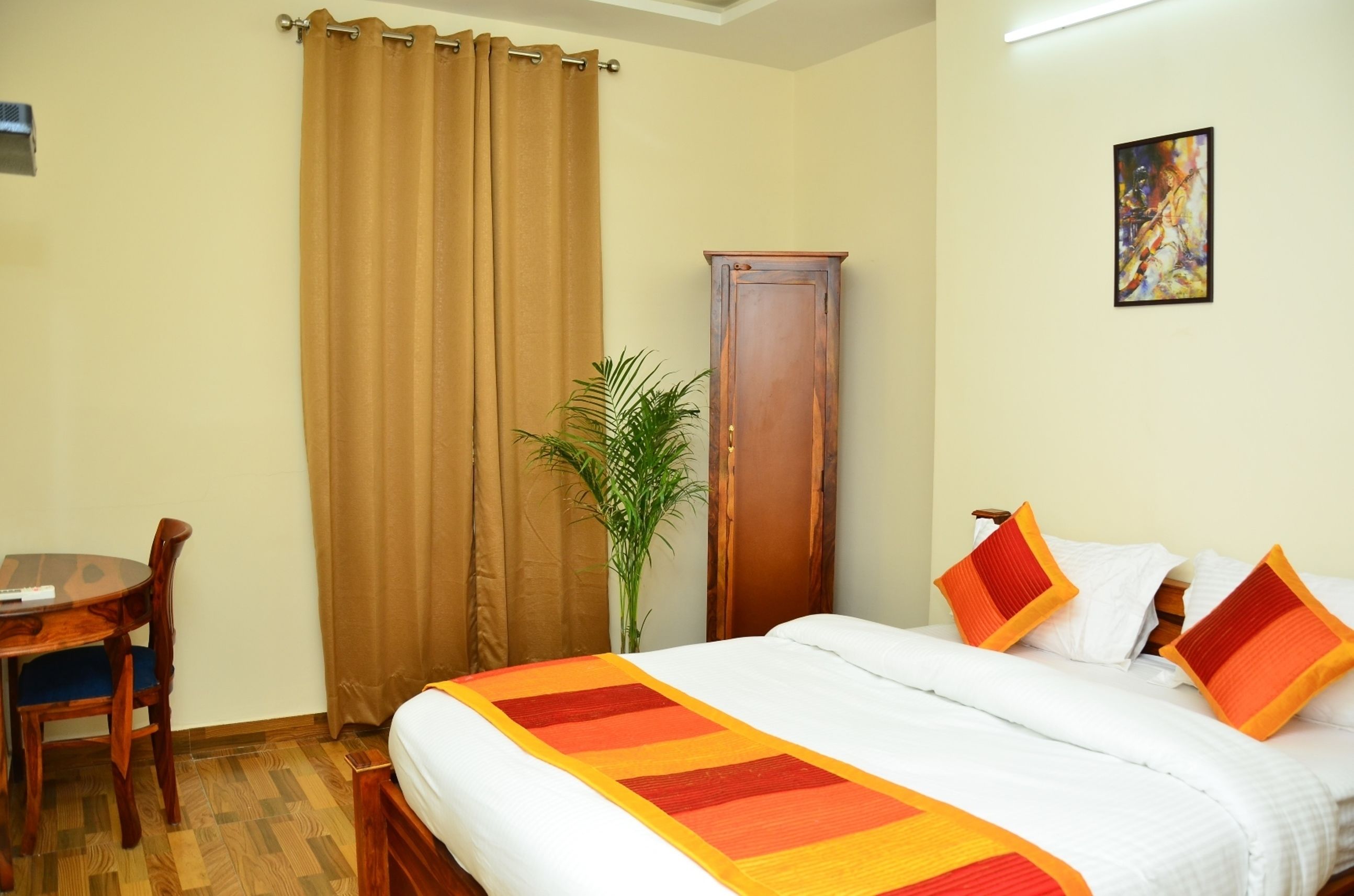Habitación de hotel con cama, mesita de noche de madera, escritorio con silla, planta en maceta y un cuadro en la pared.