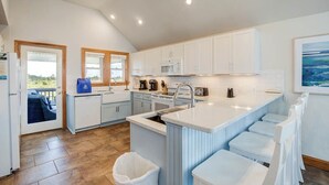 A modern kitchen with white cabinetry, a central island with bar stools, and a view through the door to an outdoor area. - Just For You | 988 ft from the beach | Dog Friendly, Private Pool, Hot Tub | Nags Head (Nags Head)