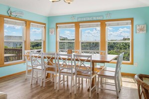Un espace repas avec une table en bois et des chaises blanches, de grandes fenêtres donnant sur un paysage verdoyant et un luminaire au plafond.