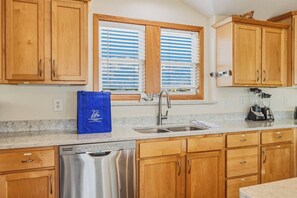 Una cocina con armarios de madera, un lavavajillas de acero inoxidable, un lavamanos con grifo y una ventana con persianas.
