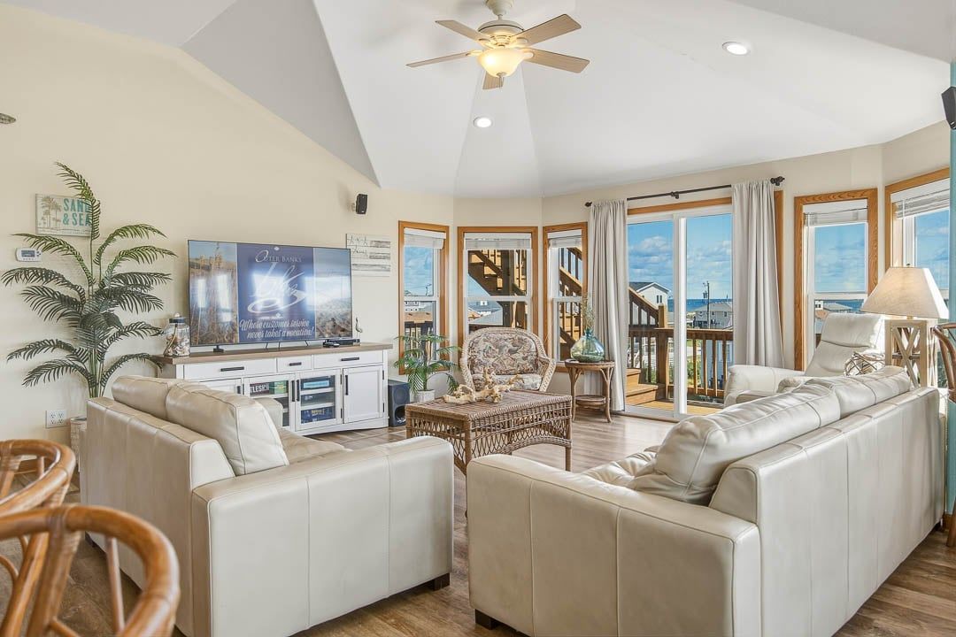 A spacious living room with a large sofa, a television, and a view of the sea through large windows.