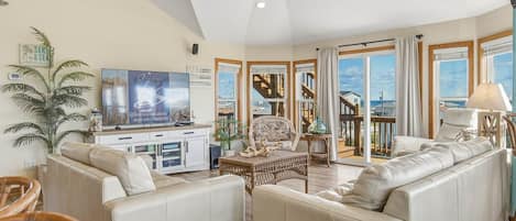 A spacious living room with a large sofa, a television, and a view of the sea through large windows.