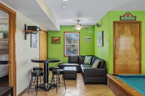 A room with a pool table, a sofa, a dining table with stools, a ceiling fan, and a door labeled 'Beach House'.