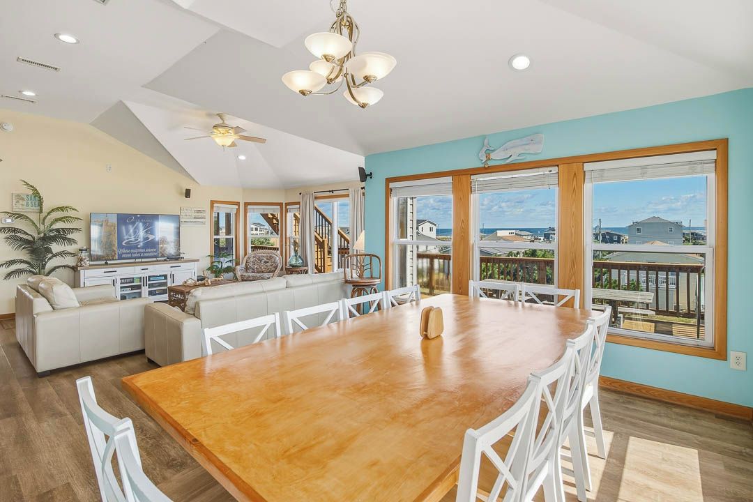 A spacious living area with a large wooden dining table, white chairs, a sofa, and a view of the ocean through large windows.