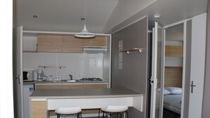 Een kleine keuken met een centraal eiland, twee barkrukken, en een bed dat zichtbaar is in de aangrenzende kamer.