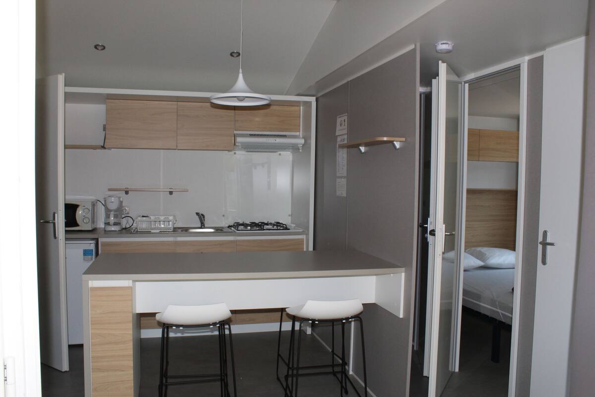 A compact kitchen with a central island, two bar stools, and a visible bed in the adjacent room.