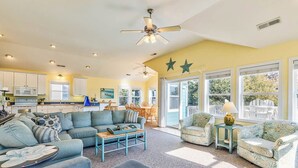 A spacious living room with a large sofa, a coffee table, and a dining area with chairs. There is a kitchen in the background and large windows providing natural light. - Bama Breeze | Sound Side | Private Pool, Hot Tub | Kitty Hawk (Kitty Hawk)