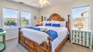 A bedroom with a wooden bed, large windows, a ceiling fan, and a decorative cabinet. - Bama Breeze | Sound Side | Private Pool, Hot Tub | Kitty Hawk (Kitty Hawk)