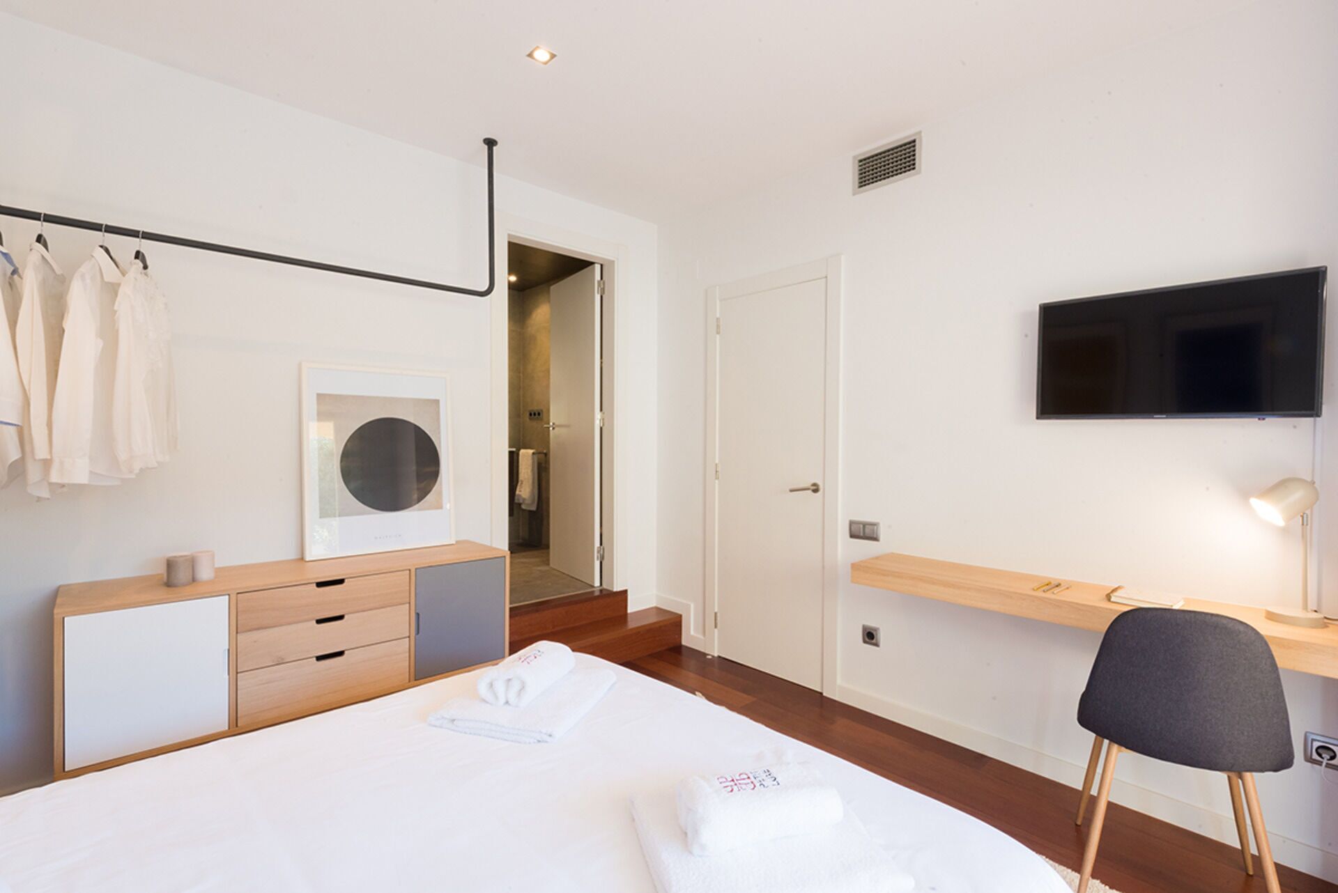 A modern hotel room with a bed, a desk, a chair, a TV, and a bathroom visible through an open door.