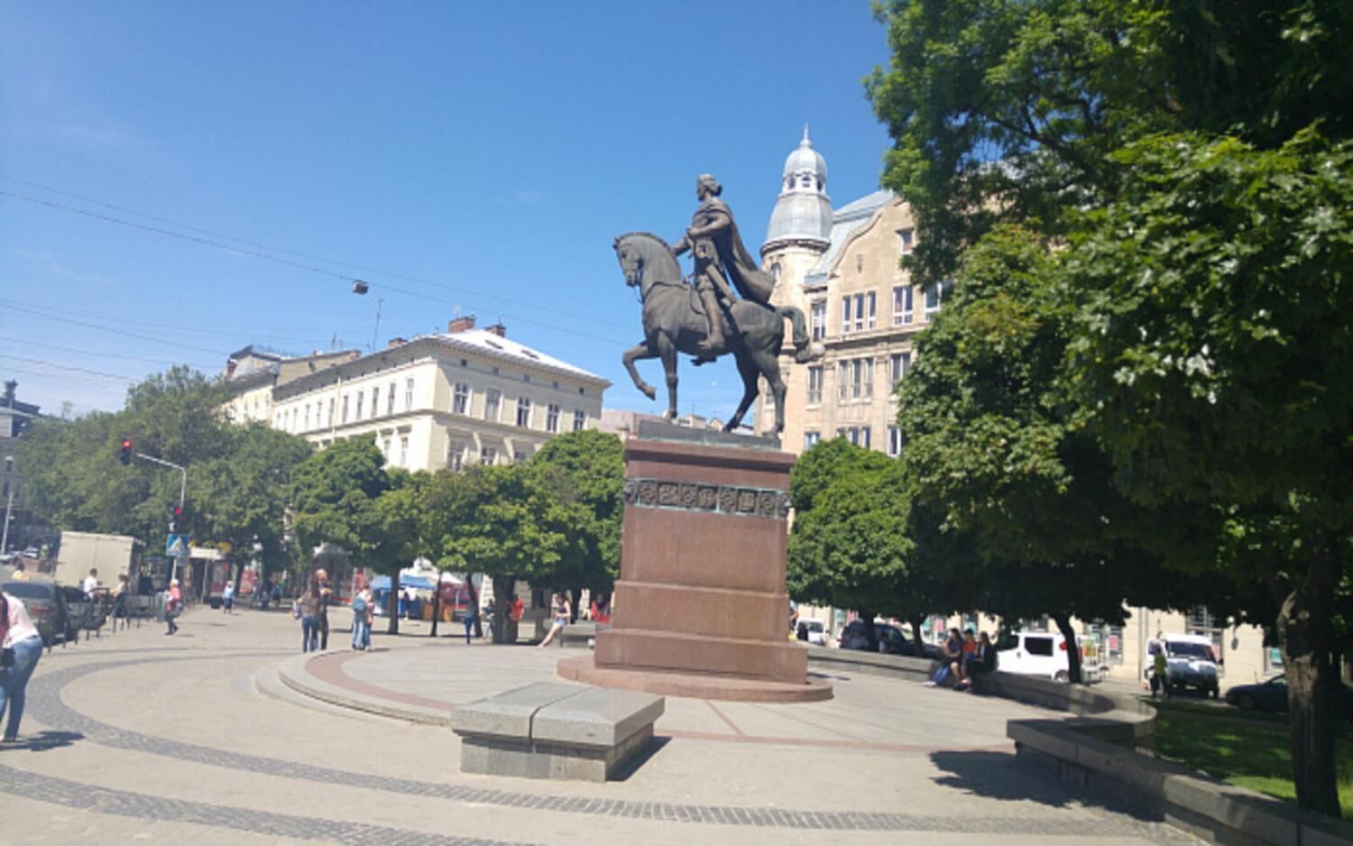 A public square with a central monument, surrounded by trees and buildings.