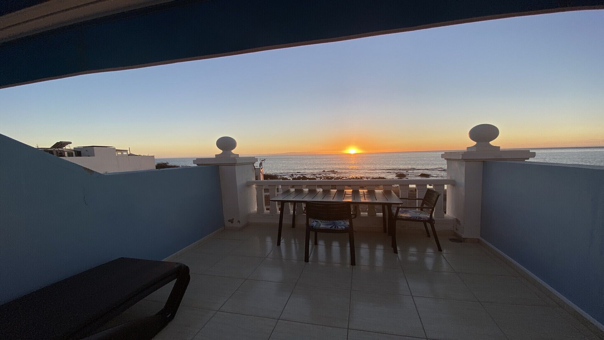 Un balcón con vista a la puesta de sol sobre el océano, con una mesa y sillas.
