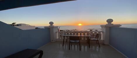Un balcón con vista a la puesta de sol sobre el océano, con una mesa y sillas.