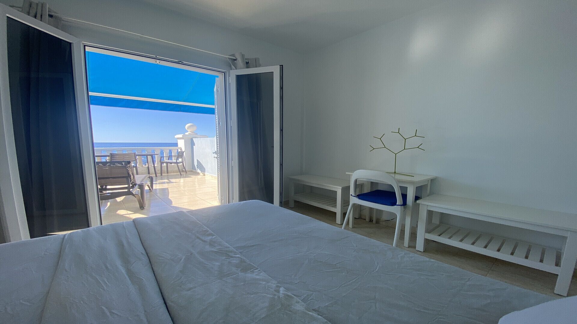 A hotel room with a bed, a desk, and a chair. There is a view of the beach through an open door.