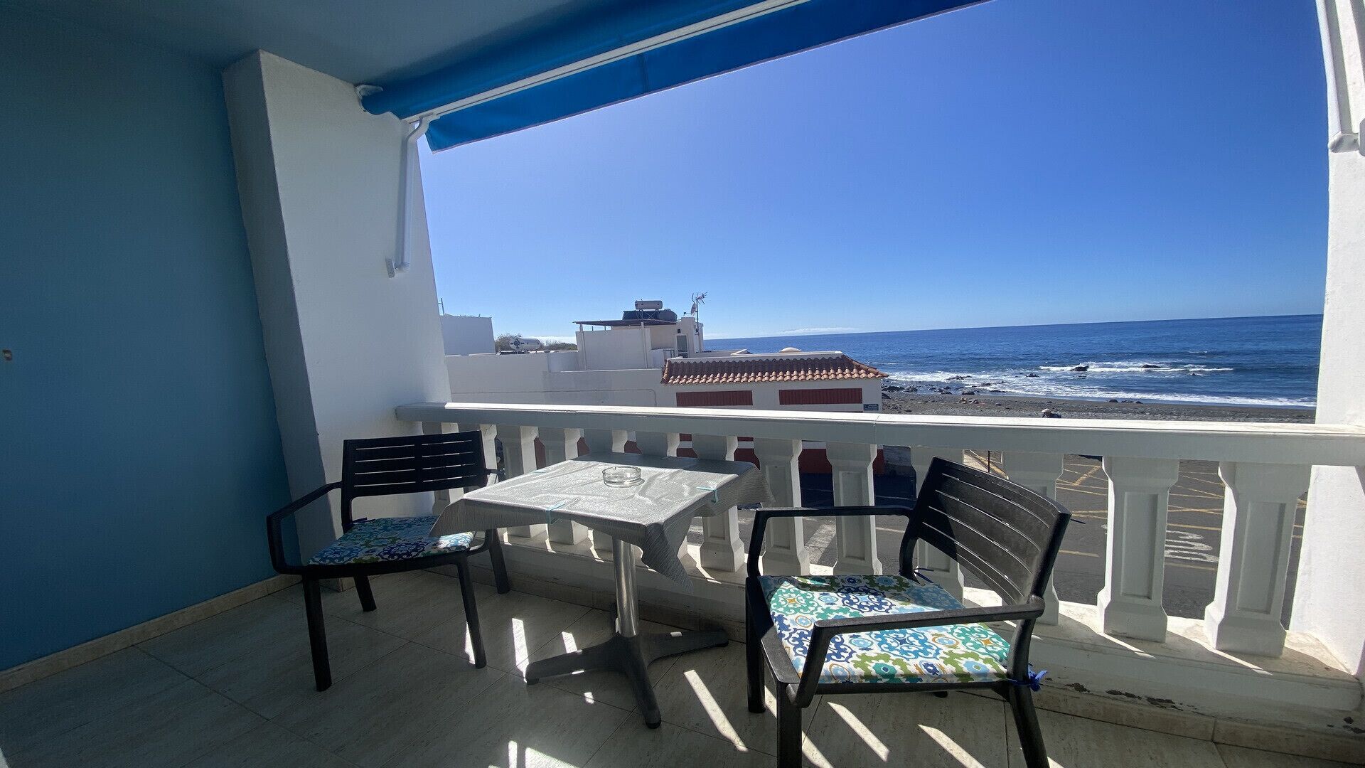 Un balcó amb dues cadires i una taula, amb vista a una platja i un edifici amb teulada vermella.