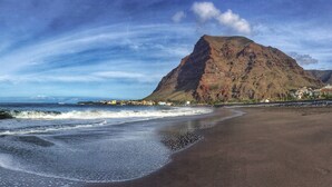A beach with waves, a mountain, and buildings in the background. - Molino azul 1A (Valle Gran Rey)