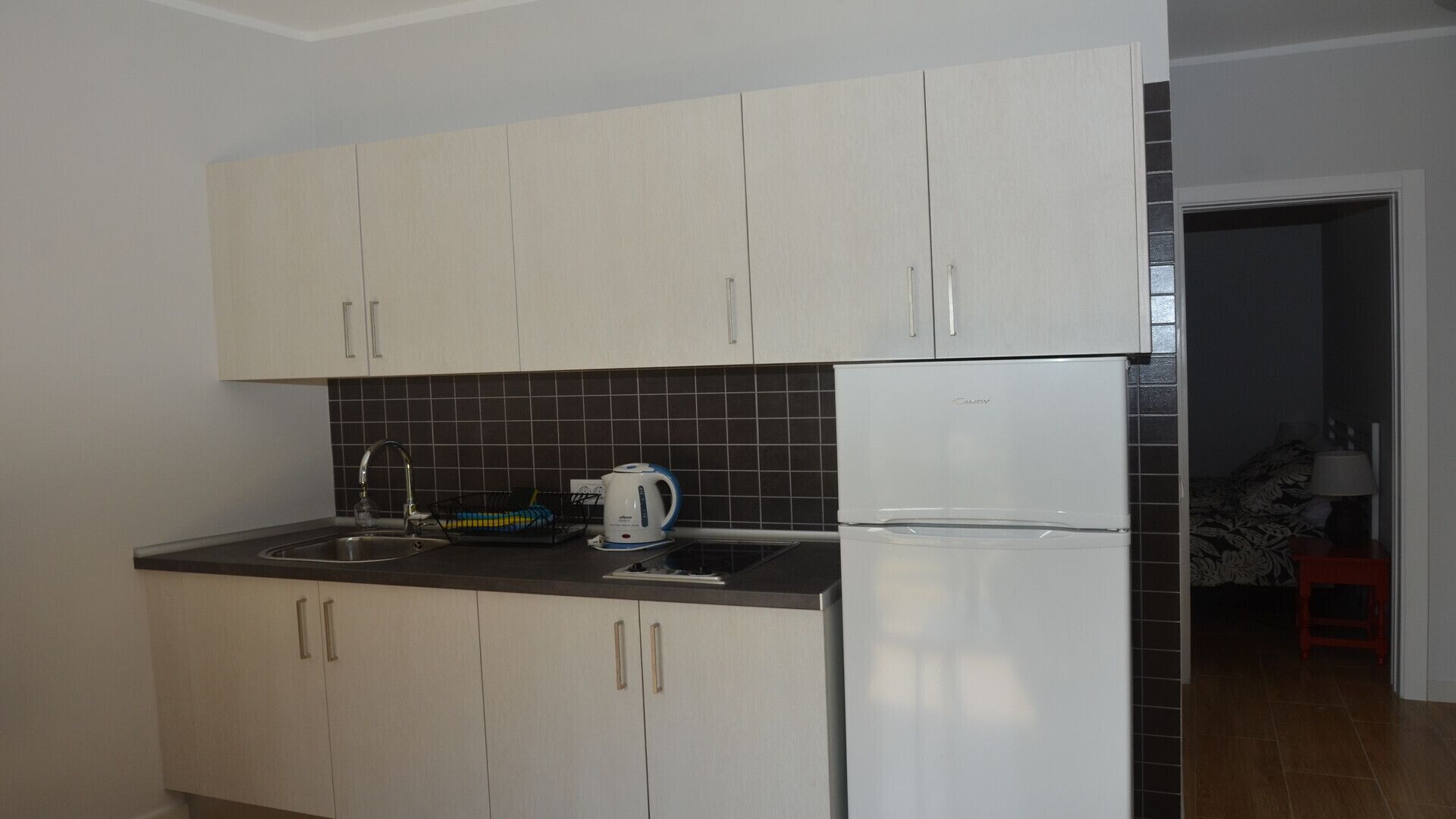 A kitchen with white cabinets, a black tiled backsplash, a stainless steel sink, a kettle, and a white refrigerator.