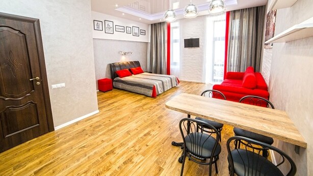 A modern living room with a wooden floor, a dining table with chairs, a sofa with red cushions, and a bed with a striped blanket. - Apartment Mitskevicha 5a (Lviv)