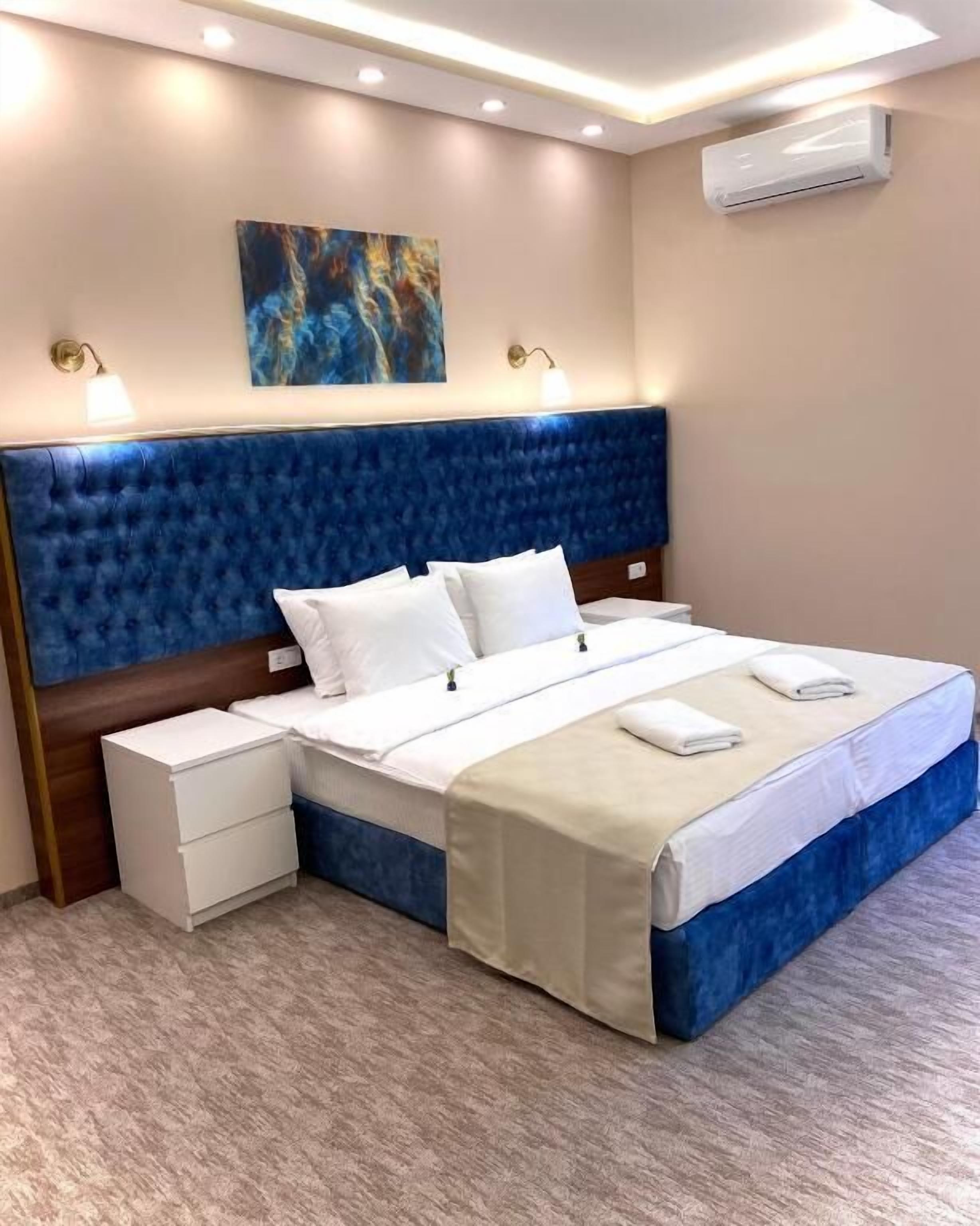 Una habitación de hotel con un cabecero tapizado en azul, una cama blanca con colcha color beige, una mesita de noche con cajón y un equipo de aire acondicionado en la pared.