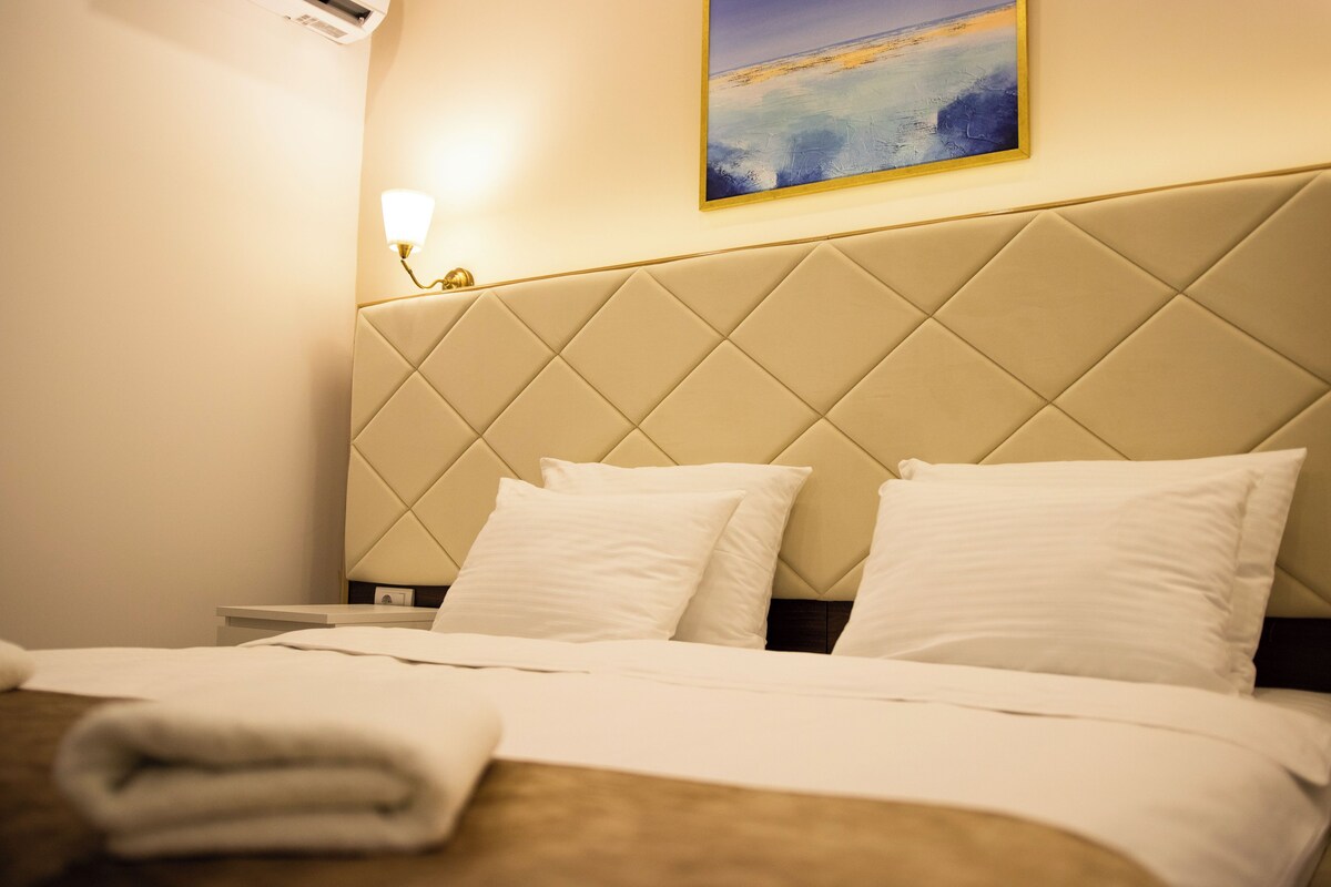 Une chambre d’hôtel avec un grand lit, du linge de lit blanc, une tête de lit à motifs matelassés, une table de chevet et une lampe fixée au mur.