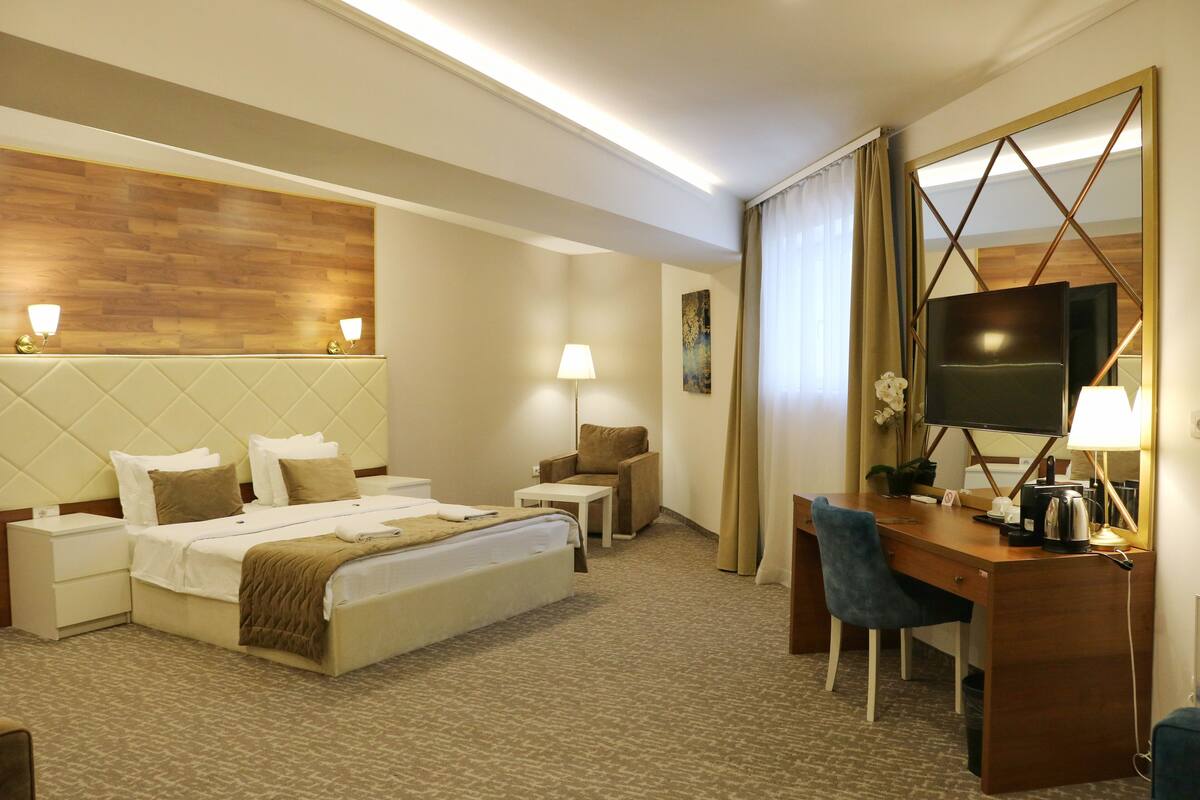 Una habitación de hotel con cama, mesitas de noche, un escritorio con silla, televisor y un espejo grande.