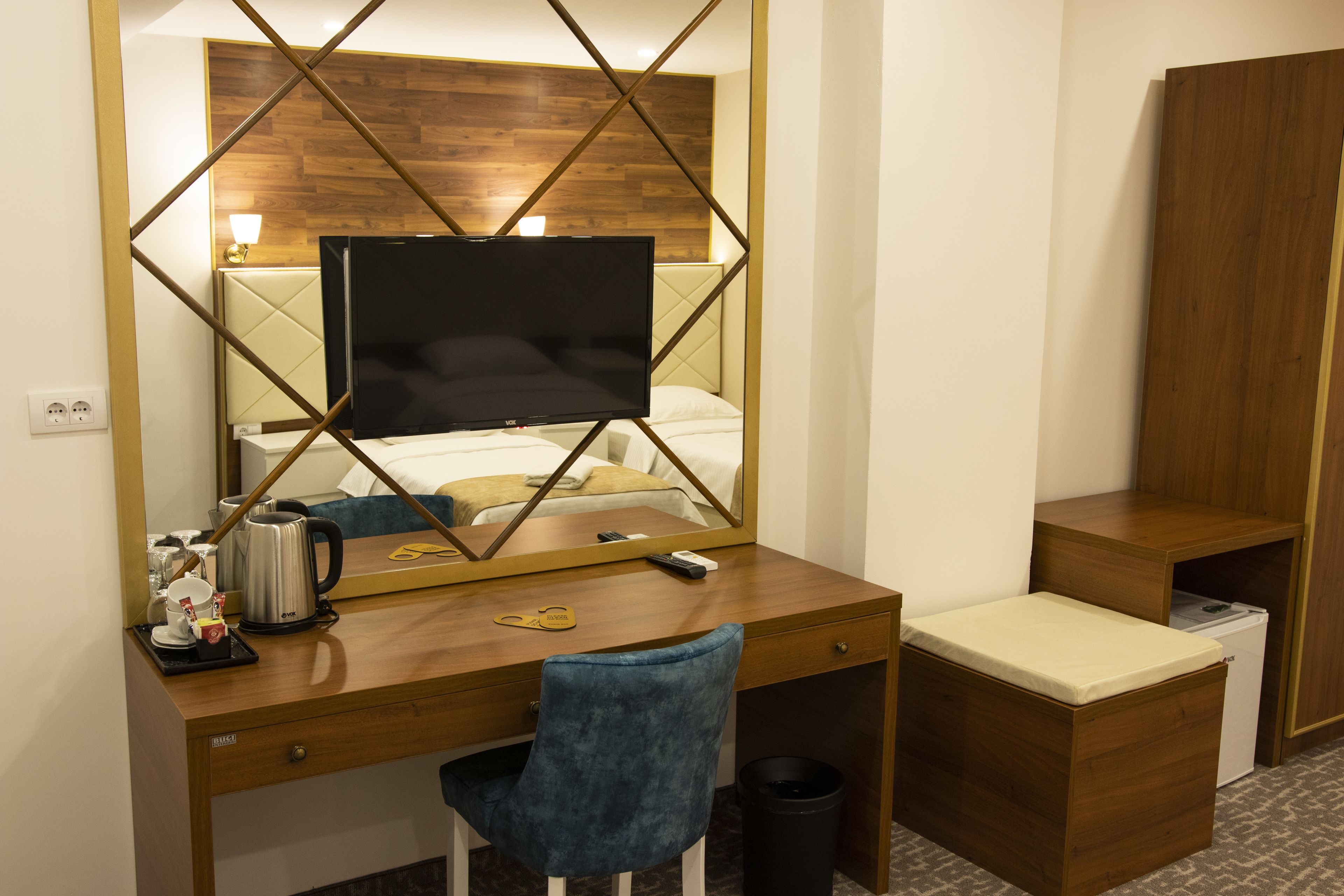 A hotel room with a wooden desk, a television, a bed with a headboard, a chair, a small trash bin, and a wardrobe.