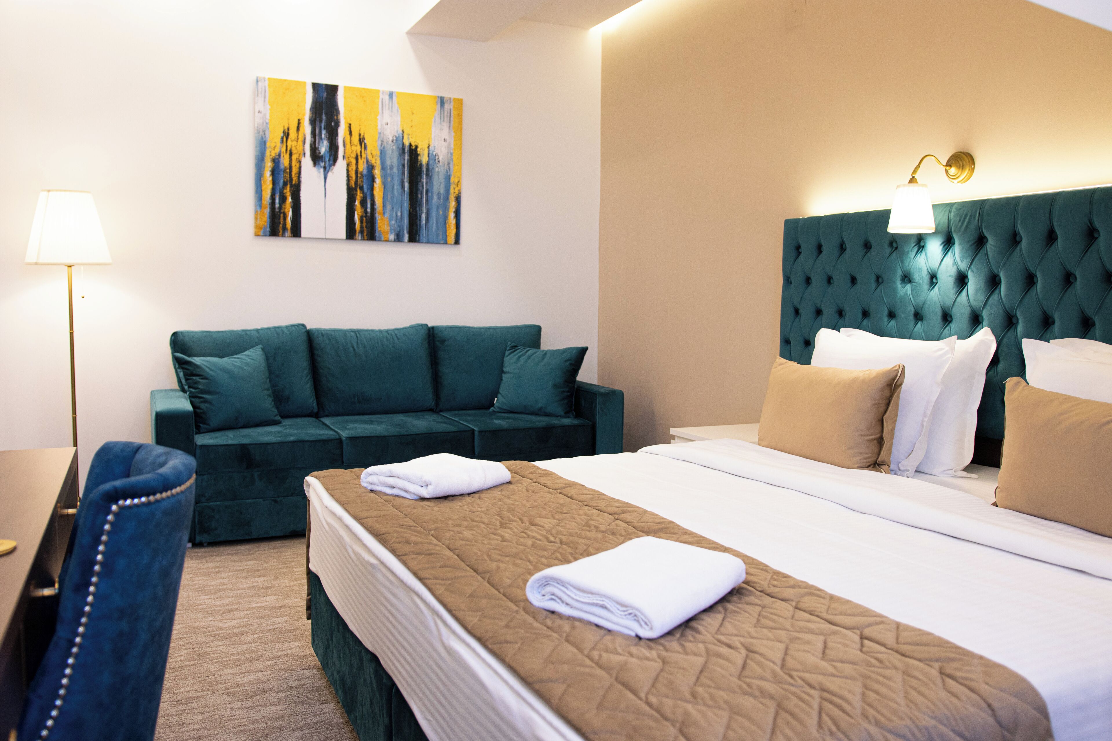 A hotel room with a bed, a green sofa, a desk with a lamp, and a painting on the wall.