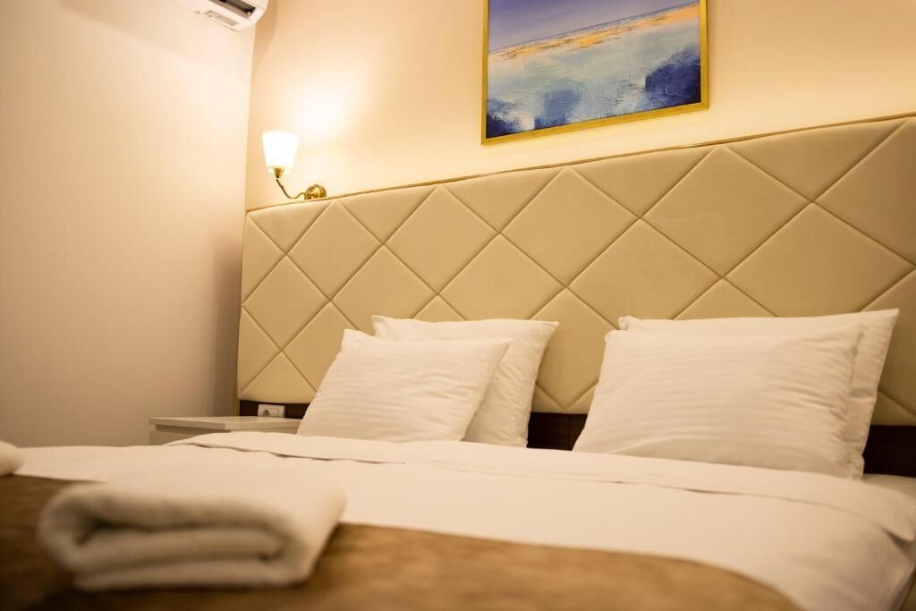 Una habitación de hotel con cabecera acolchada, cama con ropa blanca y una toalla doblada sobre la cama, además de una lámpara de pared.