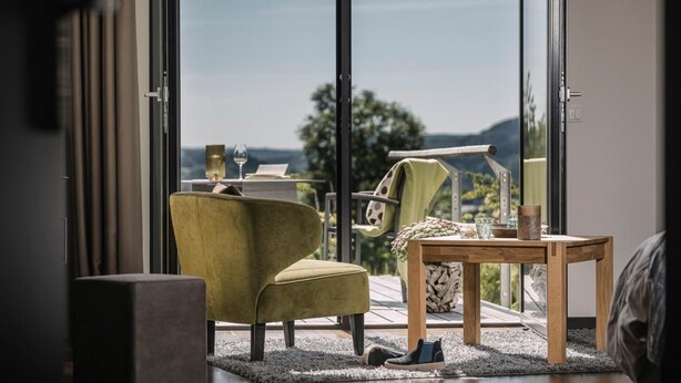 A modern room with a large window, a green armchair, a wooden table, and a view of the outdoors. - Genusswerk Eifel - Boutique Hotel (Korperich)