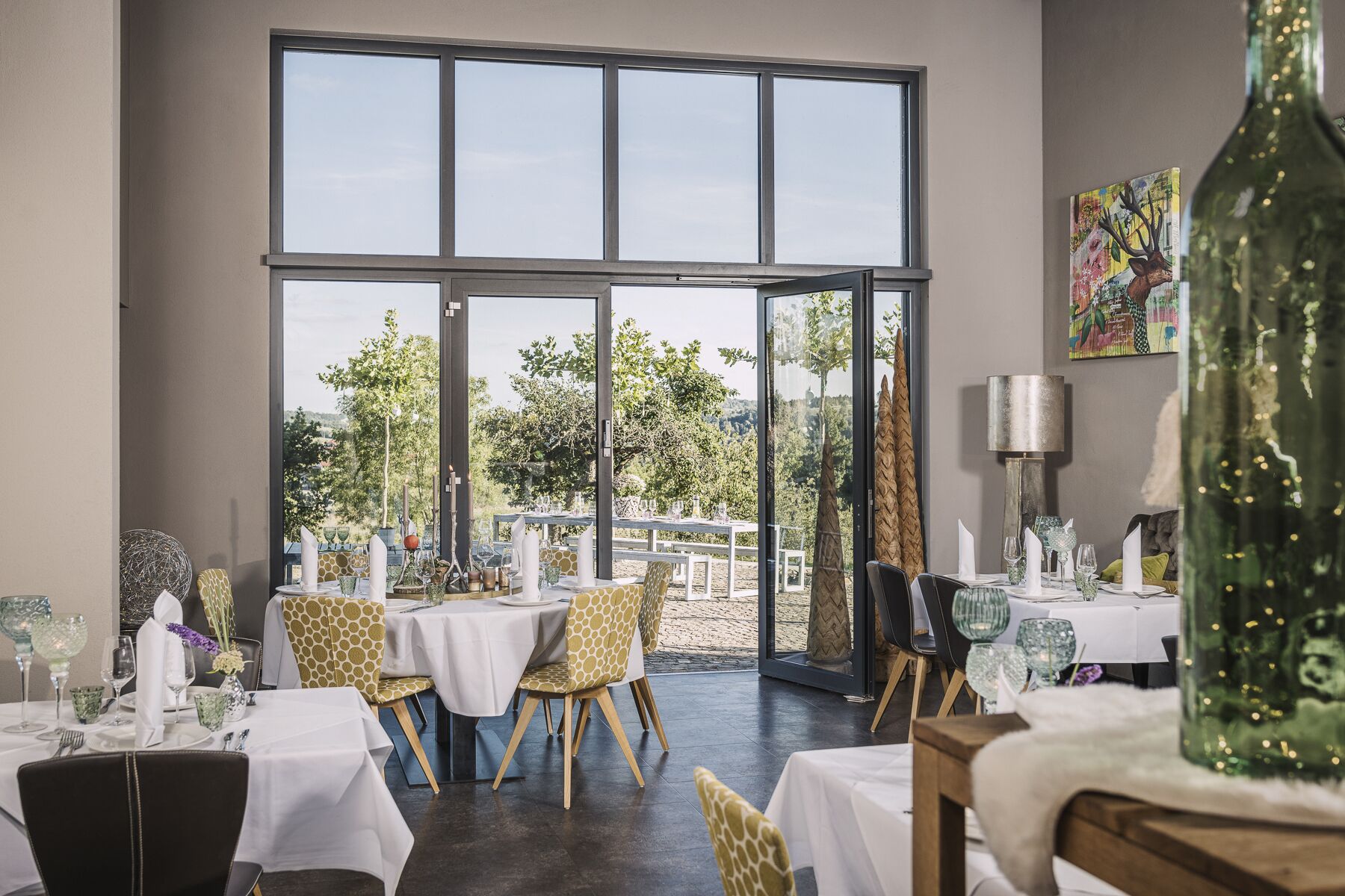 A dining area with tables set for two, large windows, and a view of the outdoors.