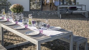 An outdoor dining area with tables set for a meal, a building entrance with a sign, and a view into the interior. - Genusswerk Eifel - Boutique Hotel (Korperich)
