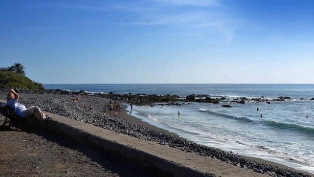 A beach with people walking and swimming, a rocky shoreline, and a clear blue sky.