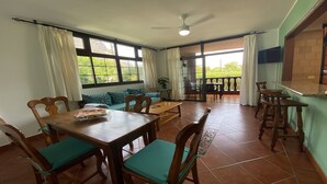 A dining area with a wooden table and chairs, a sofa, and a balcony with a view of greenery.