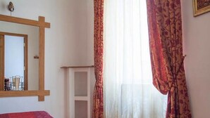 Un dormitorio con cama, espejo, ventana con cortinas y un estante blanco pequeño.