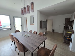 Een moderne eetruimte met een houten tafel en stoelen, een keuken met een inbouwoven, en een uitzicht door een raam.