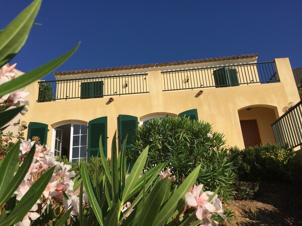 Un immeuble de deux étages avec des volets verts, un balcon et un jardin fleuri.