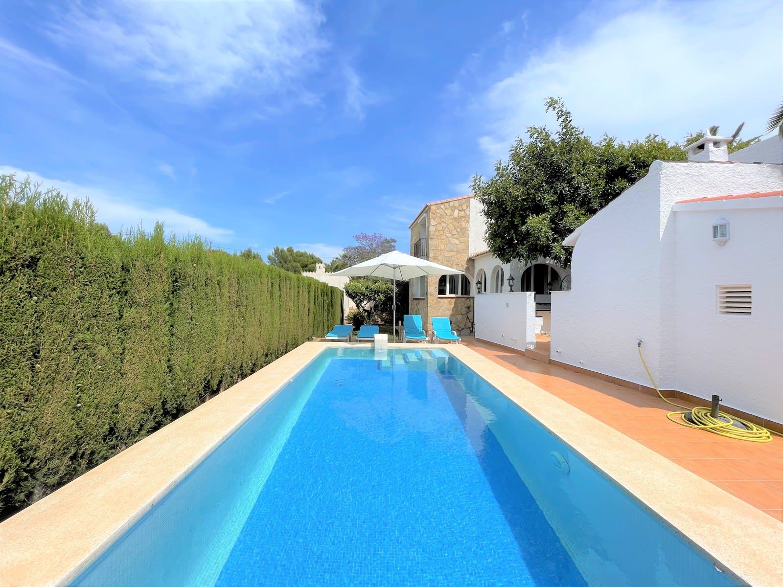 A swimming pool with lounge chairs and umbrellas, surrounded by a white building and a hedge.