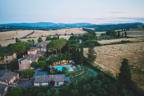Vue aérienne d’une propriété rurale dotée d’une piscine, de chaises longues et de bâtiments traditionnels en pierre, le tout entouré de champs et d’arbres.