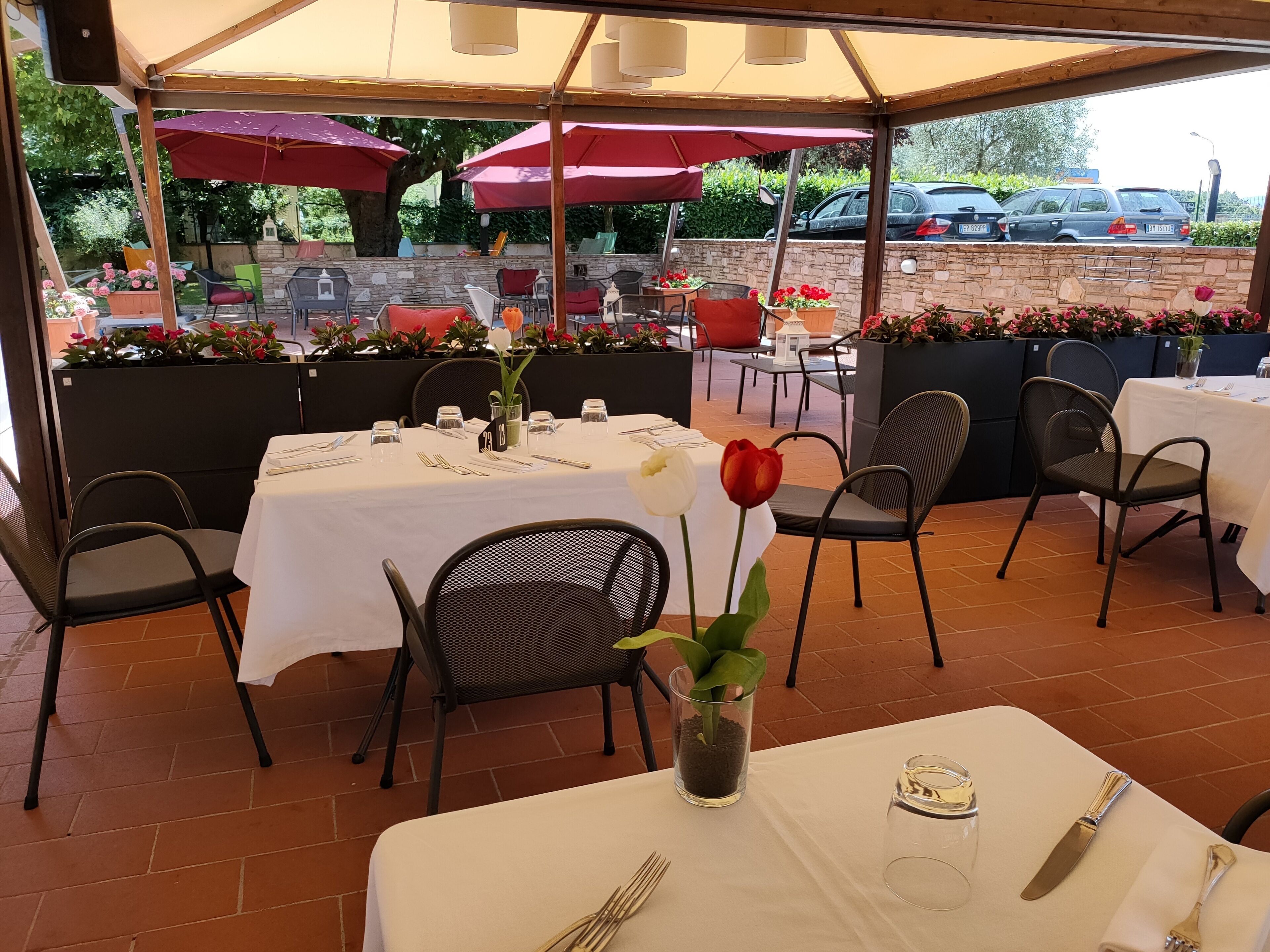 Un área de comedor al aire libre con mesas preparadas para dos, cubierta por una marquesina y rodeada de sillas y plantas en macetas.