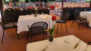 Un área de comedor al aire libre con mesas preparadas para dos, cubierta por una marquesina y rodeada de sillas y plantas en macetas.