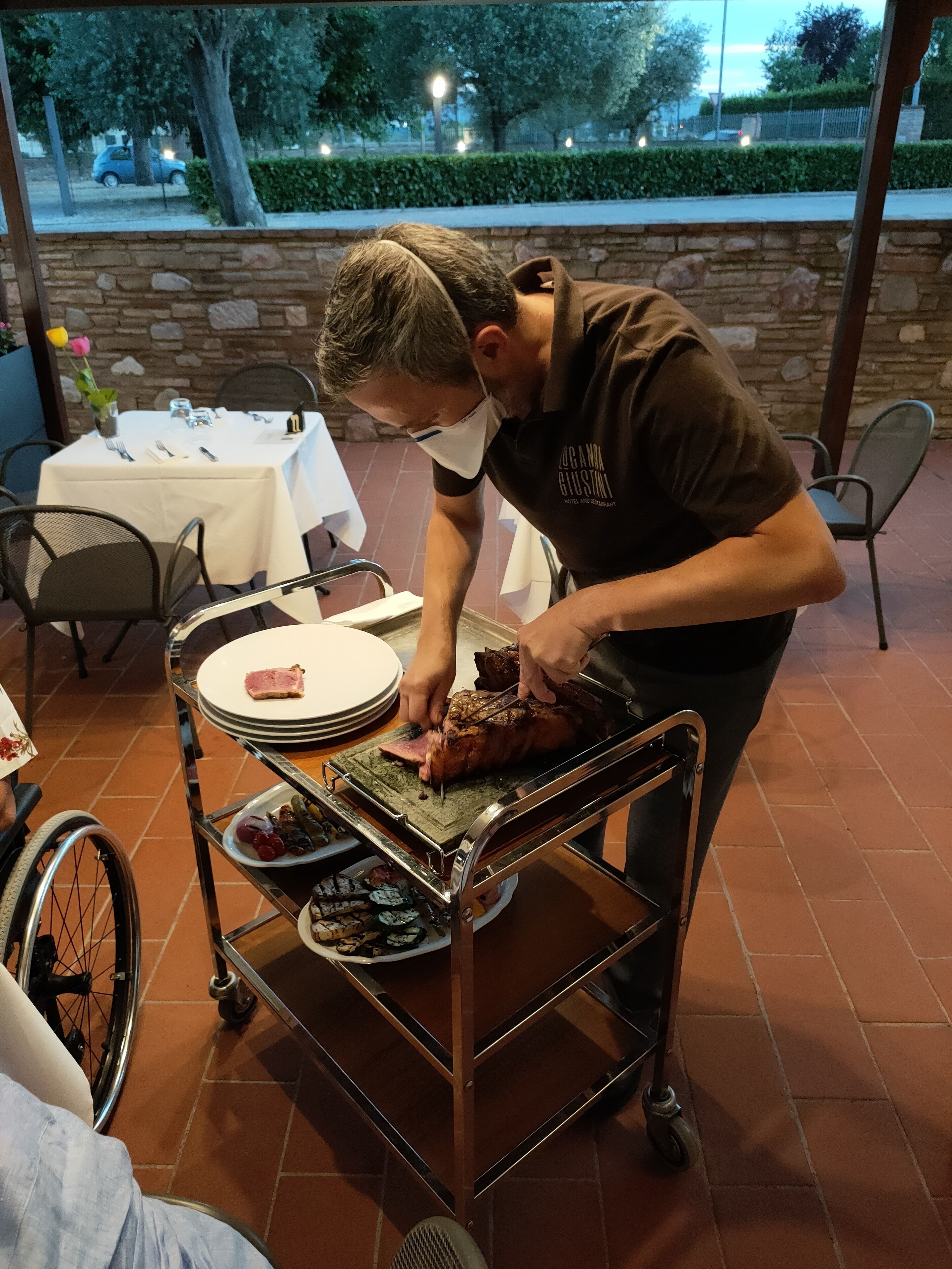 A person carving meat on a cart in an outdoor dining area with a stone wall and tables set for dining.