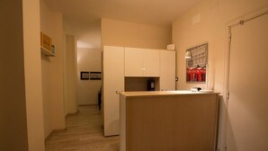 Um corredor estreito com uma mesa de recepção, uma porta à direita e um quadro na parede.