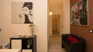 Quarto de hotel com escrivaninha, sofá preto com almofada vermelha, obra de arte na parede retratando uma mulher, e corredor que dá para outro quarto.
