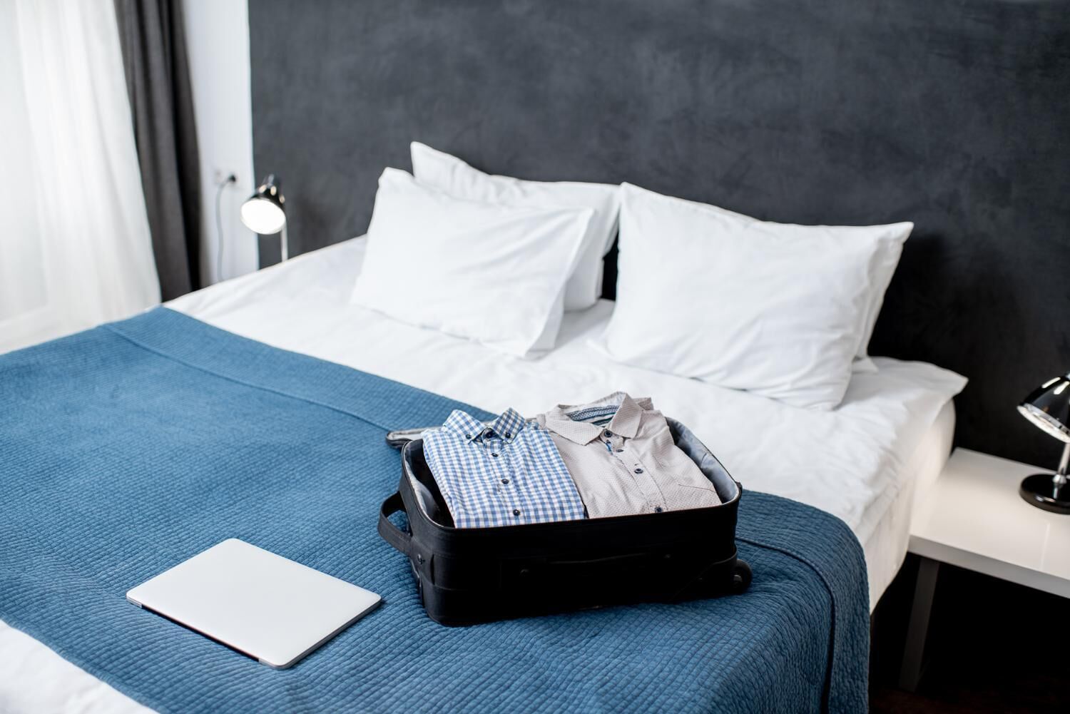 A neatly made bed with a blue blanket, a black suitcase with clothes, and a laptop on the bed.
