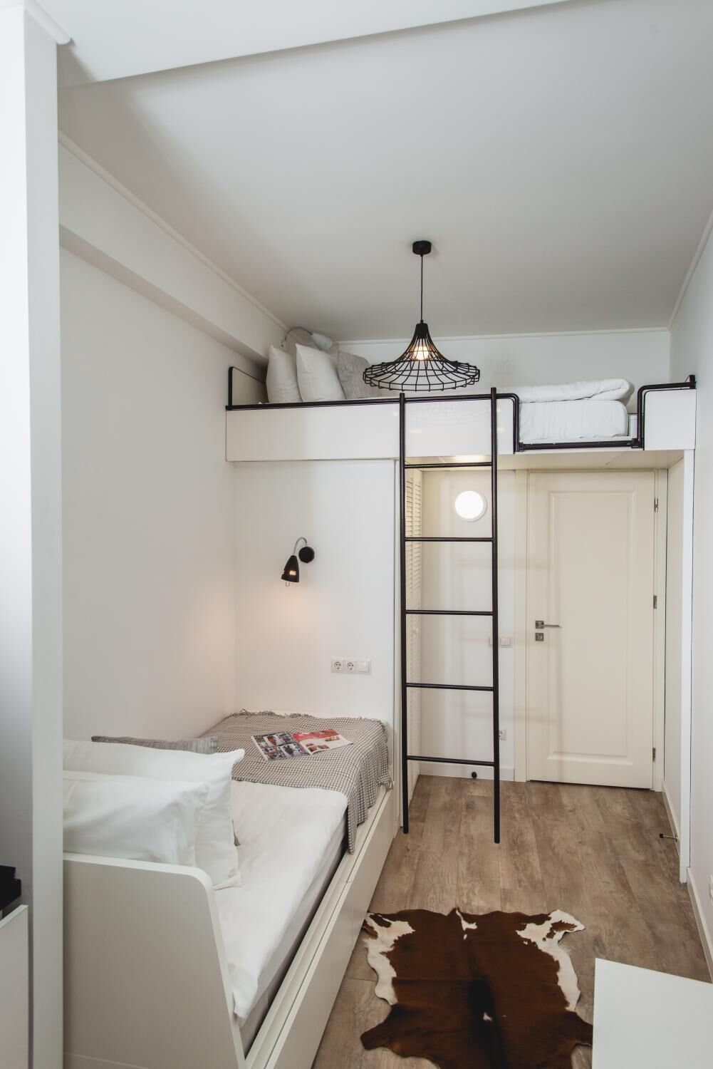 A compact bedroom with a bunk bed, a ladder, a hanging light fixture, a door, and a cowhide rug.